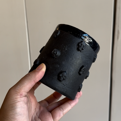 A hand holds a handleless black mug with skulls symmetrically around it in concentric rings. The top has a shiny black glaze while the body is matte black.