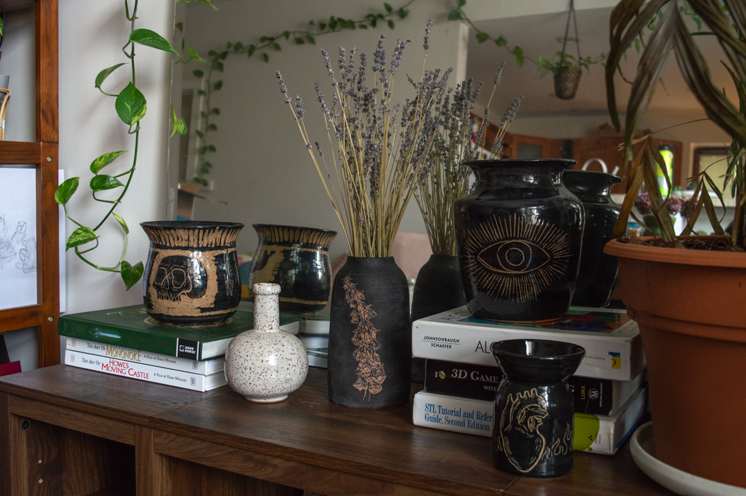 multiple ceramic pieces in black and white sit on a table with a mirror behind them. From left to right there is a large ceramic vessel with a skull, a small circular orb bottle, a tall vase with foxglove carvings, a larger grecian vase with an eye, and a small black vase with an anatomical heart. Sitting on books like a cinematice guide to Howl's Moving Castle and Zelda. There are plants in the background.