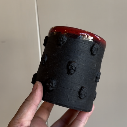 A hand holds a handleless black mug with skulls symmetrically around it in concentric rings. The top has a shiny red glaze while the body is matte black.